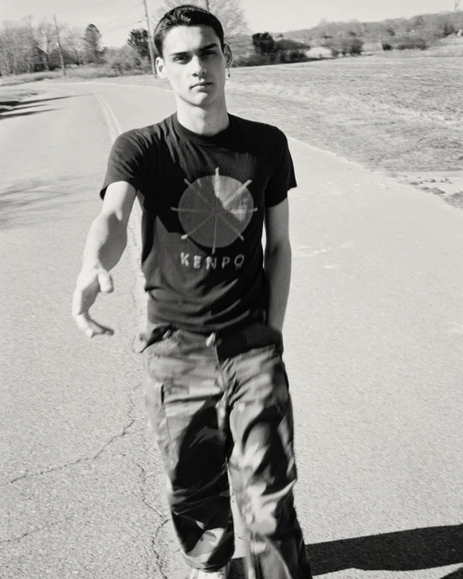 Black and white portrait of a young adult male wearing a graphic tee and camouflage pants walking outdoors on a paved road on a sunny day.