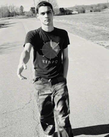 Black and white portrait of a young adult male wearing a graphic tee and camouflage pants walking outdoors on a paved road on a sunny day.