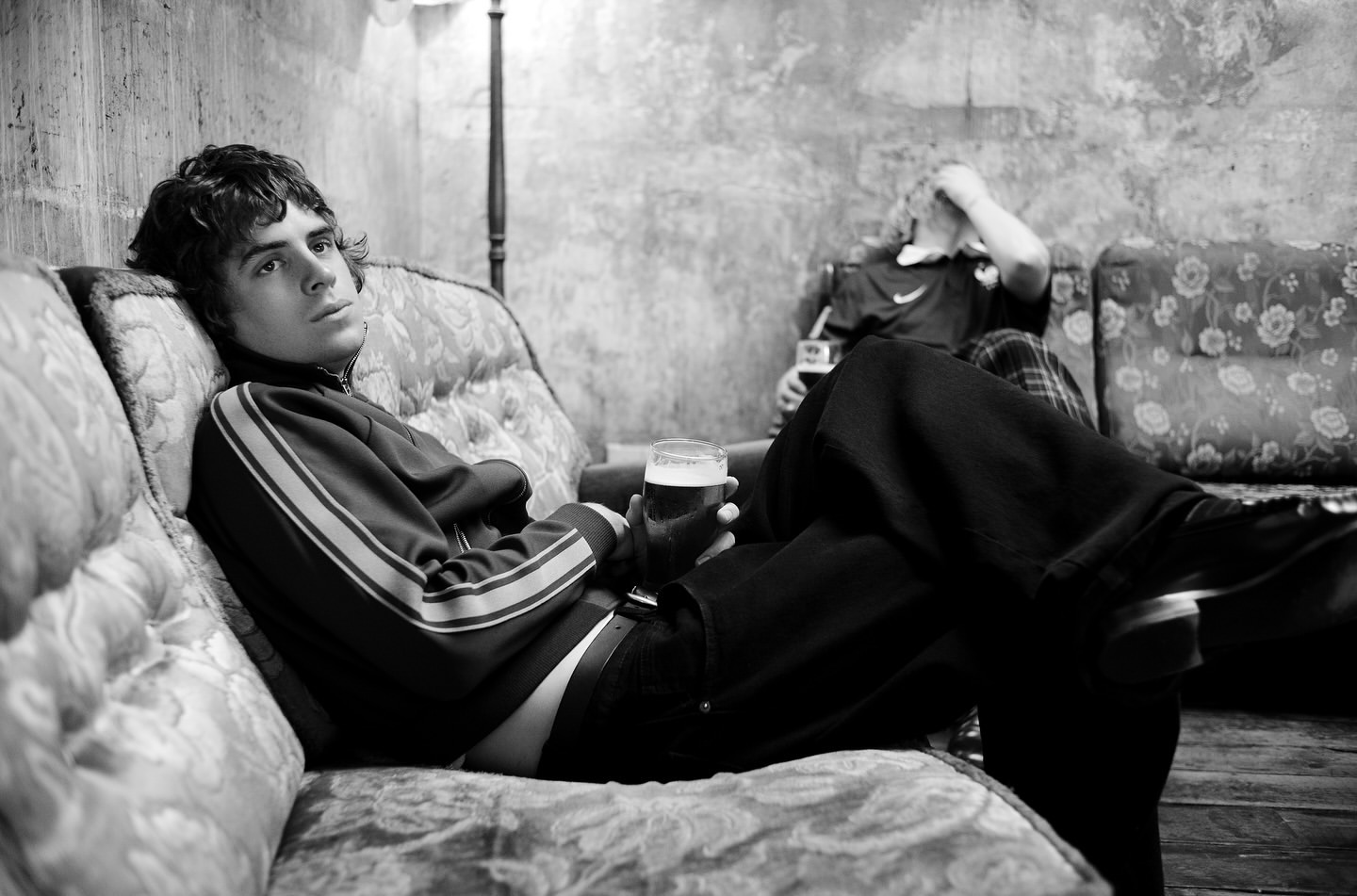 Monochrome shot of a young man in track jacket on a patterned couch with a beer, another man in the background, in a dimly lit room.