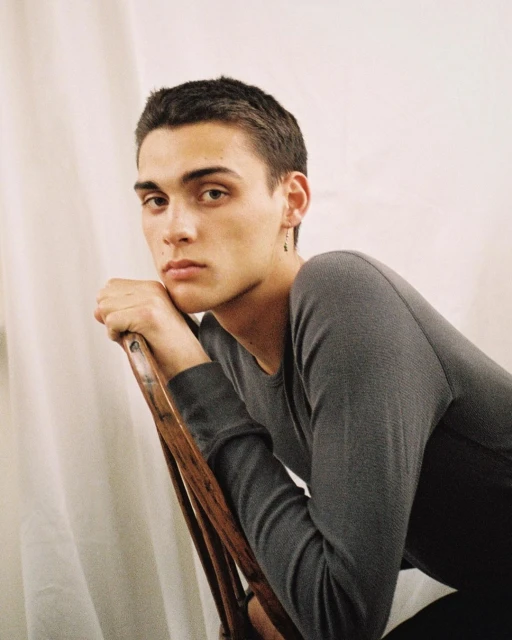 Indoor portrait of a young adult male wearing a grey long-sleeve shirt, leaning on a wooden chair against a white background.