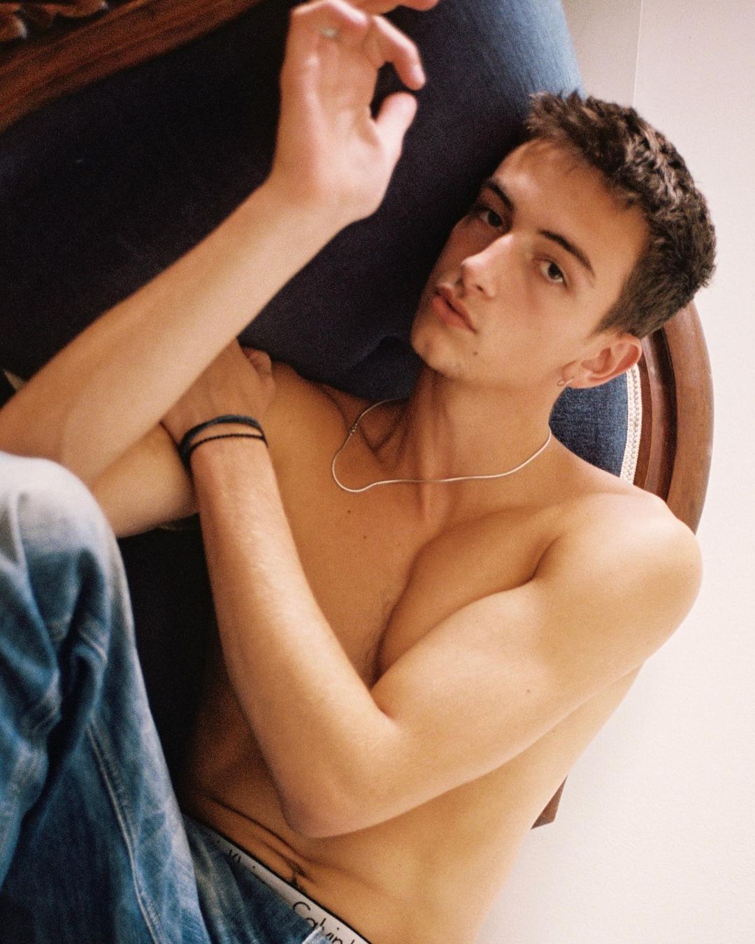 Handsome young adult male in jeans lounging shirtless against a blue cushioned chair, with fair skin, dark hair, and a silver necklace.