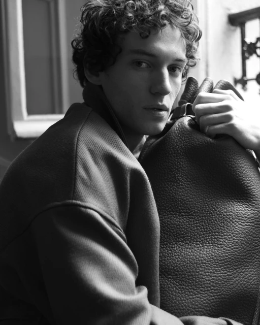 Monochrome portrait of a curly-haired young adult male in an overcoat with a bag, gazing toward the camera against a blurred indoor background.