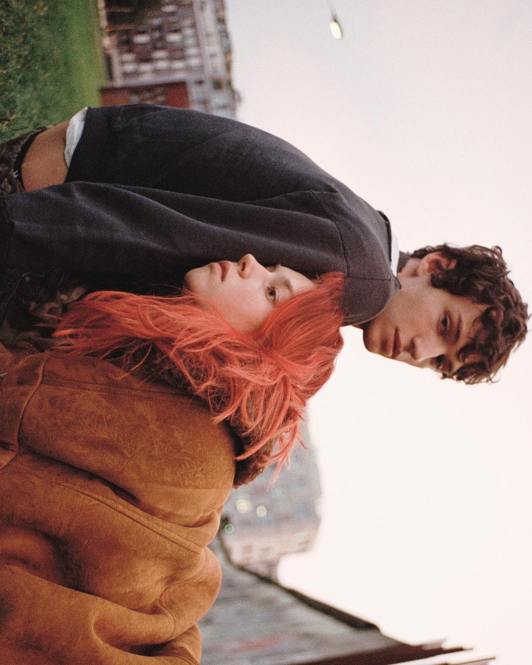 A young couple lies on the grass in an urban park, the woman with red hair, the man with dark hair, against a backdrop of buildings.