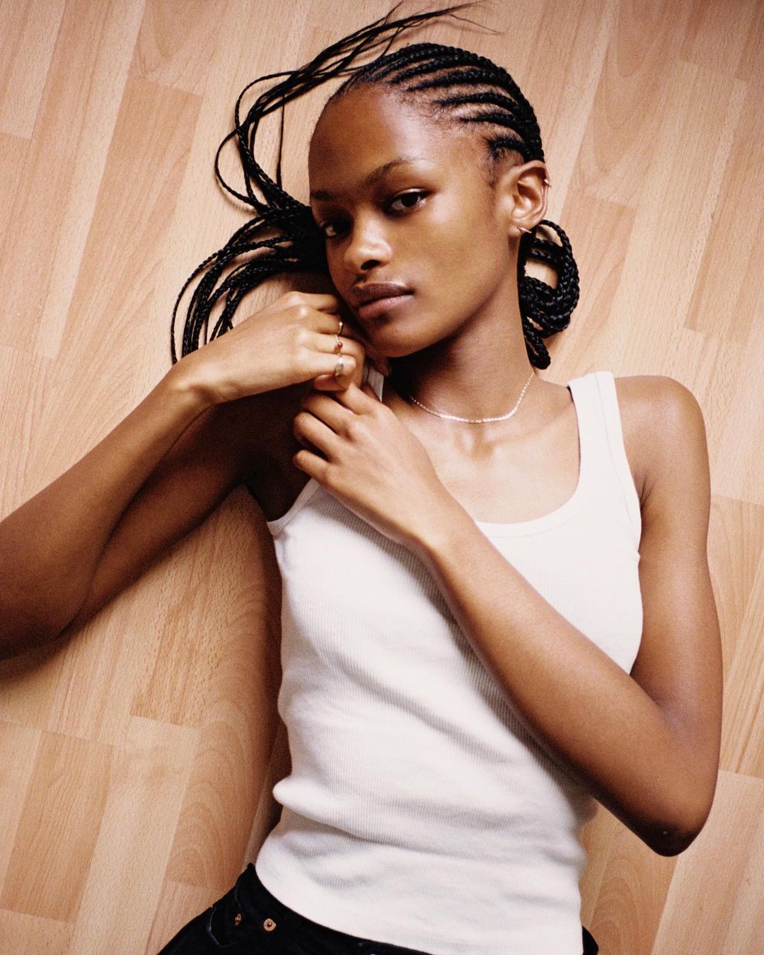 Woman with cornrow braids in a white tank top lies on a wood floor, looking at the camera with a neutral expression, indoors.