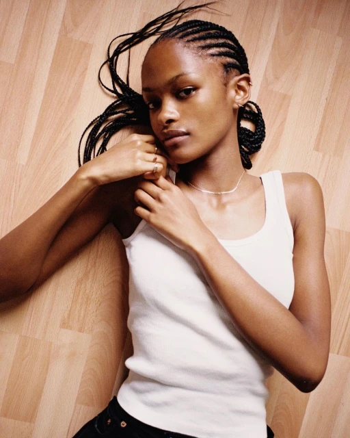 Woman with cornrow braids in a white tank top lies on a wood floor, looking at the camera with a neutral expression, indoors.