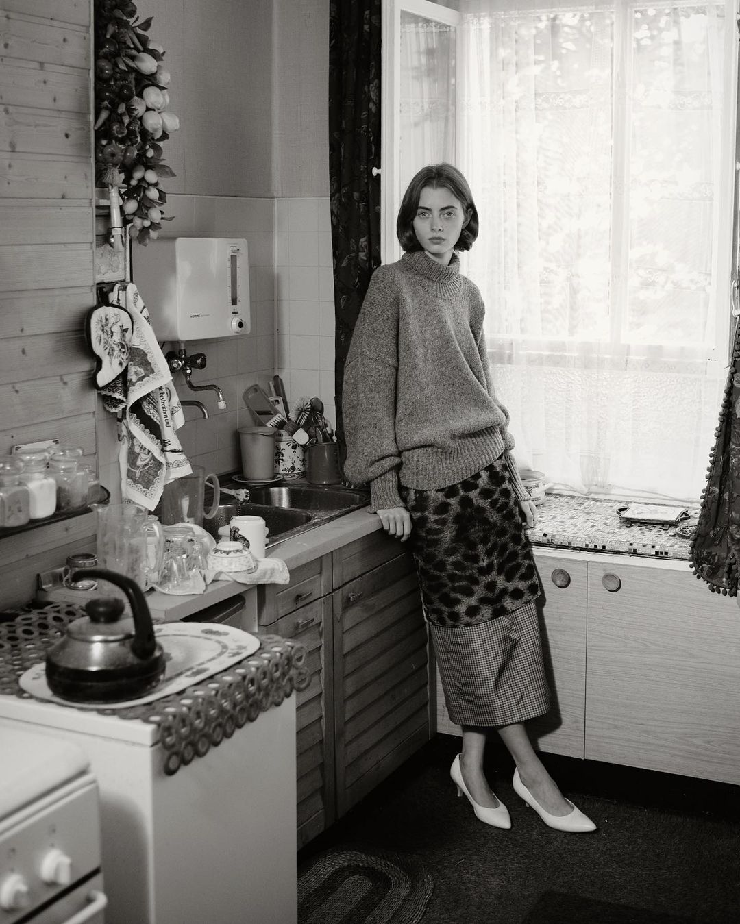 A woman leans against a kitchen counter in a black and white photograph. She wears a sweater, patterned skirt, and heels. A window provides light.
