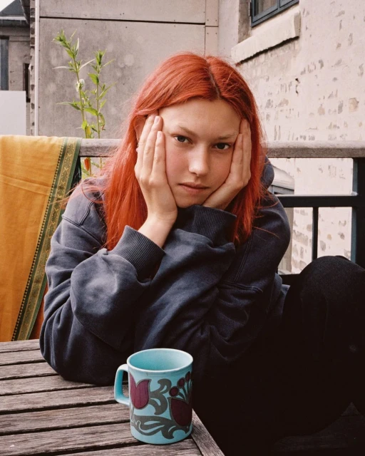 Young woman with bright red hair sits at a wooden table with a blue and maroon floral mug in front, posing with hands on her face.