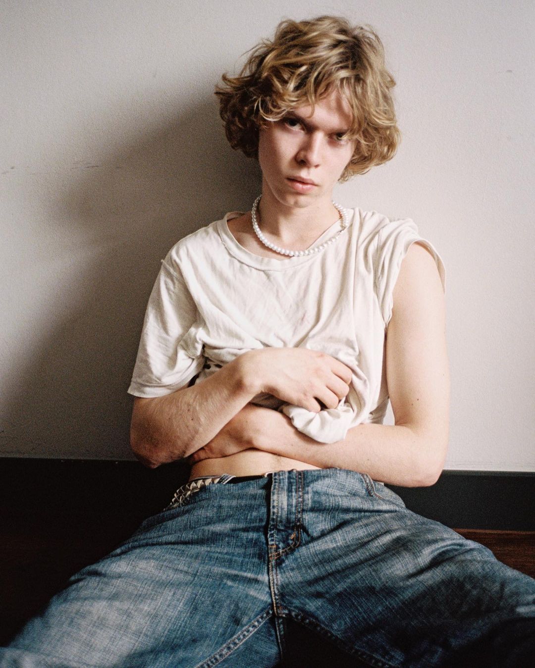 A young adult with curly blond hair poses indoors against a white wall, wearing jeans and a casual white t-shirt.