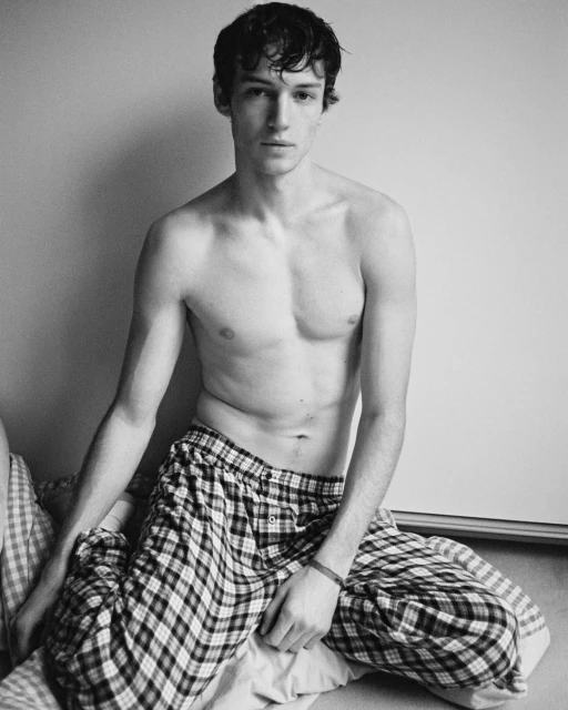 Young adult man posing indoors. He's shirtless with plaid pajama pants, sitting on a bed in soft, neutral light against a blank wall.