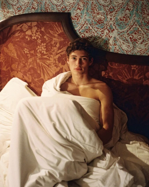 Portrait of a shirtless young adult male sitting in a bed covered by a white blanket, posing against a patterned headboard and wallpaper.
