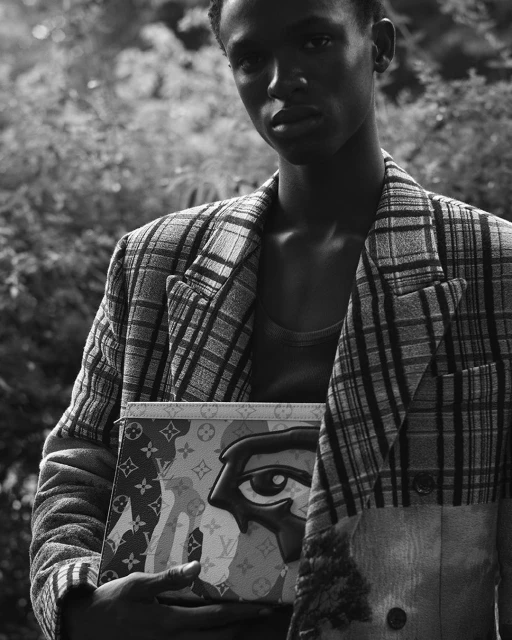 Black and white portrait of a Black male model wearing a plaid blazer holding a printed handbag outdoors in a wooded area.