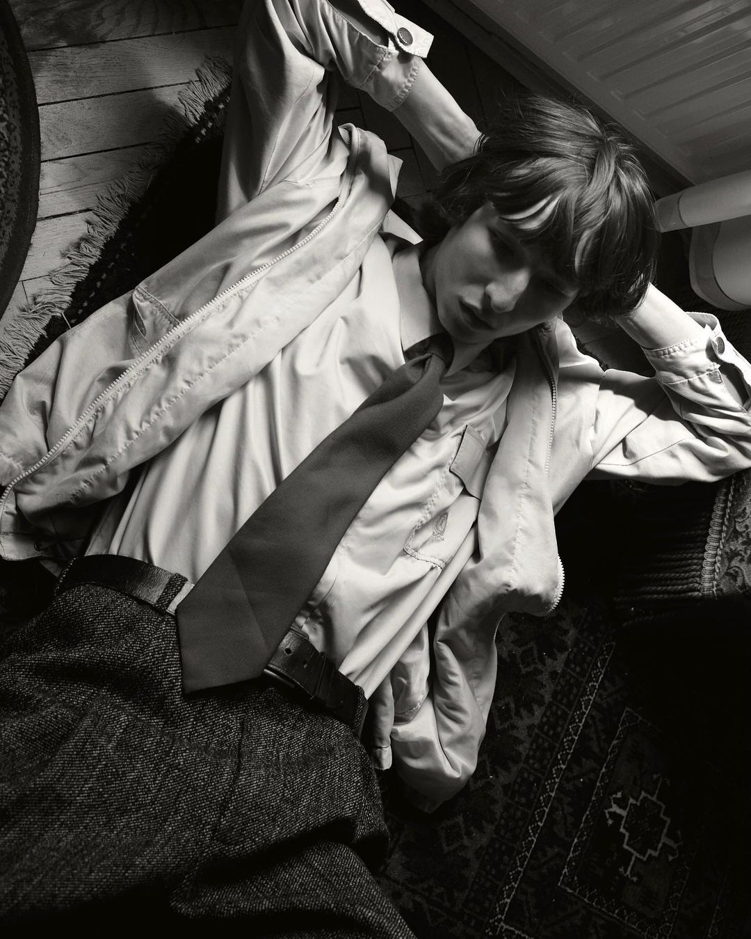 Monochromatic portrait of a young person lying on a patterned rug. The individual is wearing an unbuttoned shirt and dark tie.