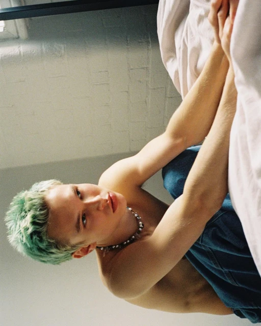 Close up, low-angle view of shirtless young male with dyed green hair wearing blue jeans and a pearl necklace posing in a light interior.
