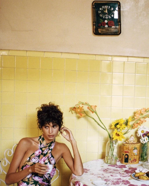 Woman in floral halter top sits at a table with tea, flowers, and cookies in a yellow-tiled room, styled with vintage clock, enjoying a quiet moment.