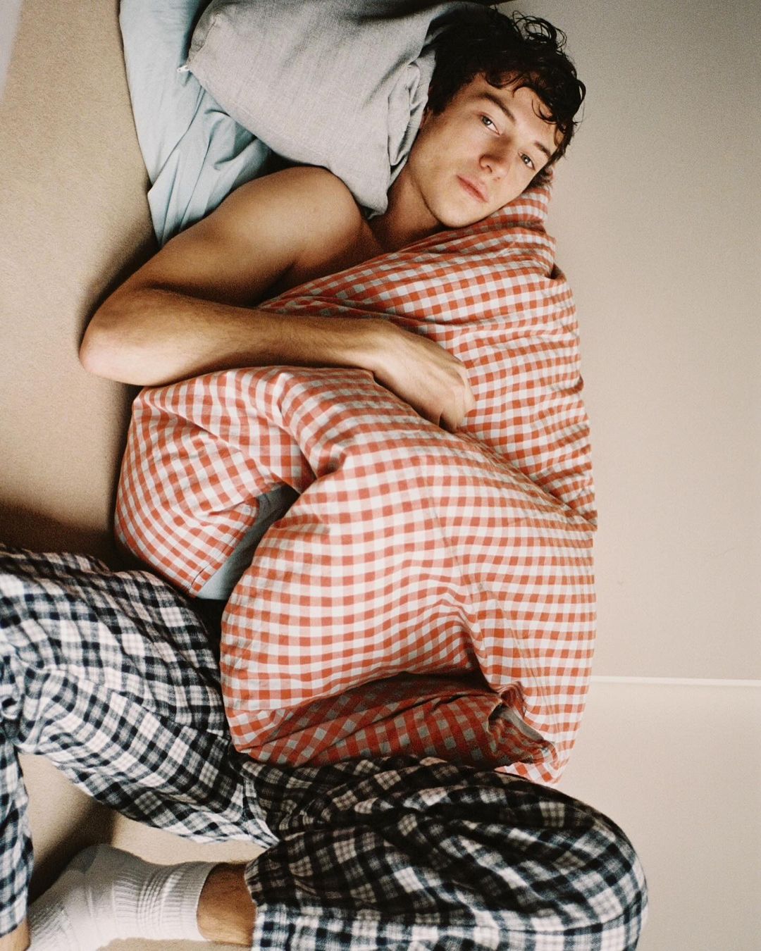 Young adult male wrapped in a red and white checkered blanket, lounging with black and white checkered pajama pants indoors.
