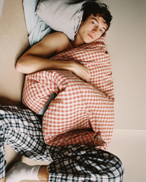 Young adult male wrapped in a red and white checkered blanket, lounging with black and white checkered pajama pants indoors.