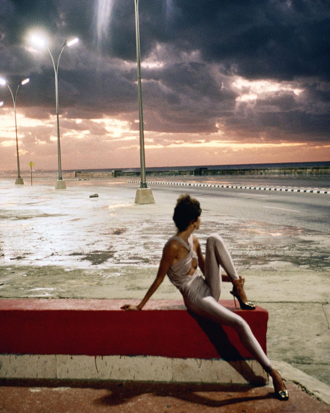 Woman in shiny bodysuit relaxes on red bench along a coastal road at dusk with streetlights under cloudy sky; fashion editorial.