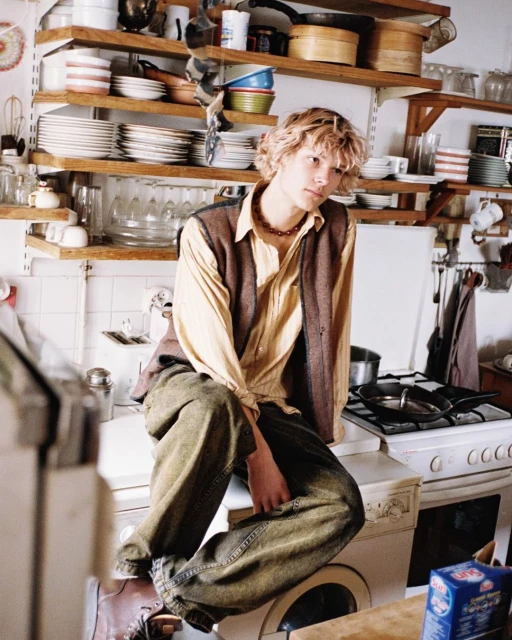 A young adult male sits on a washing machine in a cluttered kitchen with open shelves filled with dishes and cookware.