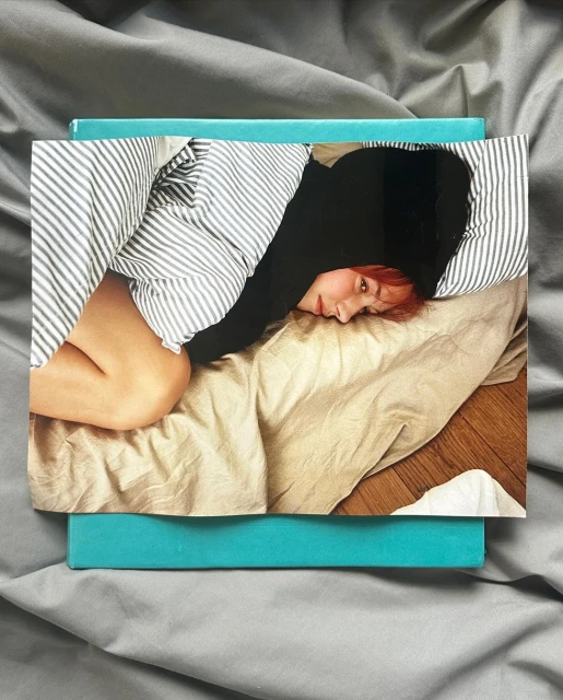 Photograph of a person lying on a tan-colored bed with striped bedding, resting on a gray sheet with a turquoise box underneath.