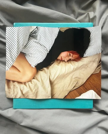Photograph of a person lying on a tan-colored bed with striped bedding, resting on a gray sheet with a turquoise box underneath.