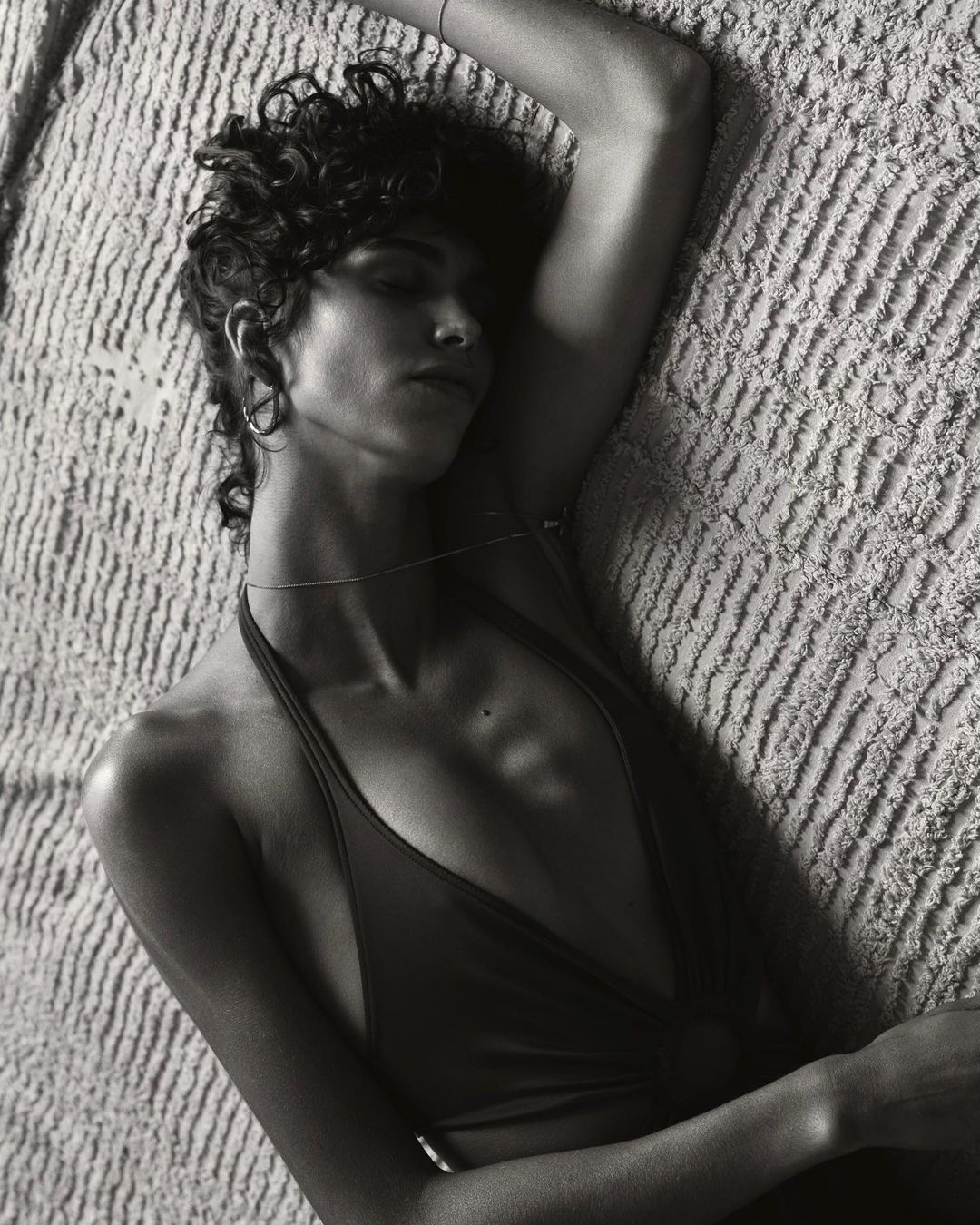 Black and white portrait of a woman with curly hair lying down, wearing a halter top, shot from above against a textured background.