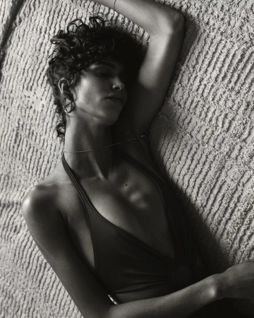 Black and white portrait of a woman with curly hair lying down, wearing a halter top, shot from above against a textured background.