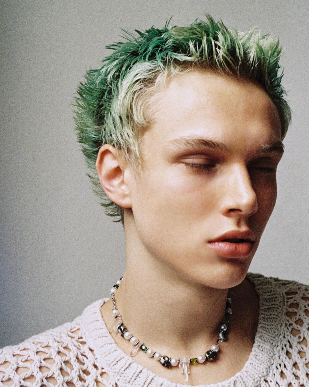 Portrait of a young adult male with green-tinted hair, eyes closed, wearing a pearl necklace and a crocheted shirt against a neutral background.