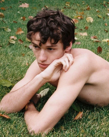 A shirtless male model with curly brown hair lies on a grassy field with scattered brown leaves in a natural outdoor setting.
