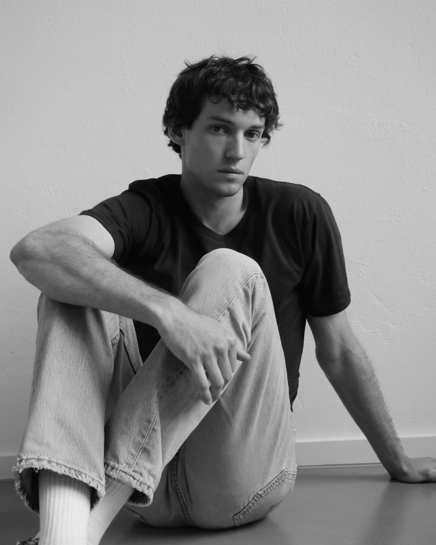 Young man in dark t-shirt and light denim jeans seated on the floor against a plain wall by Daniyel Lowden