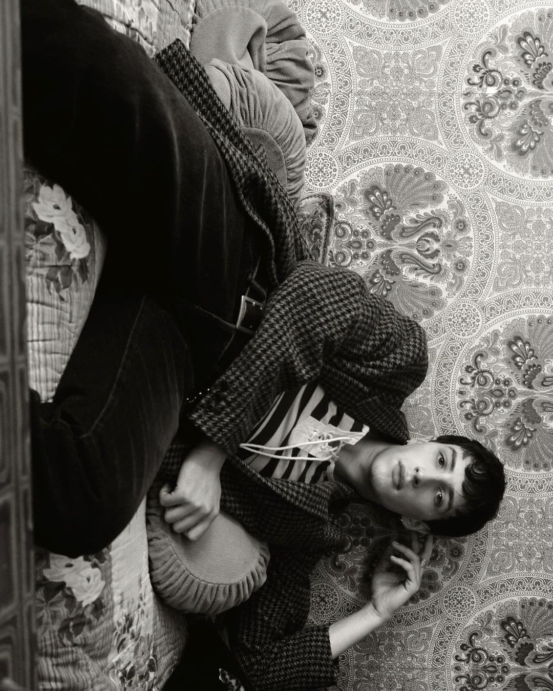A young adult lays on a patterned rug wearing a striped shirt under a checked coat, with a matching cushion and dark pants.