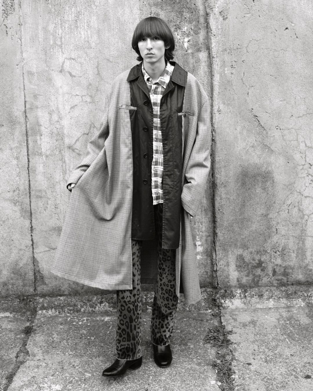 Full-length portrait of a male adult with dark hair wearing a patterned coat, shirt, and pants standing against a concrete wall in outdoor setting.