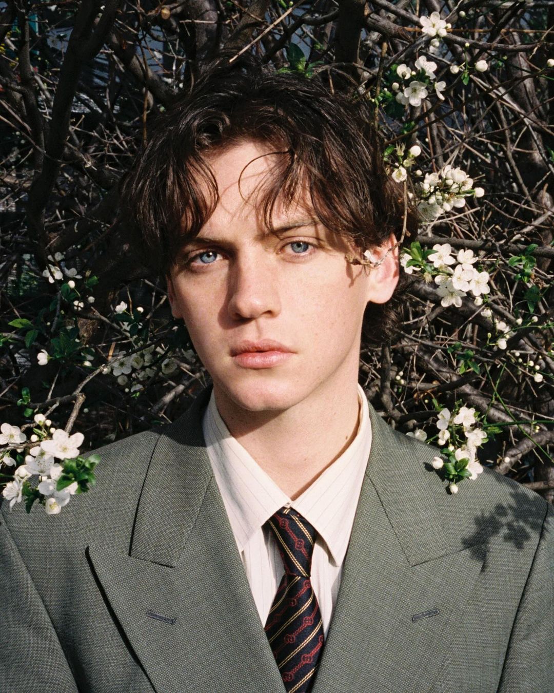 Young man in a suit and tie stands outdoors near a tree with white blossoms. The portrait features a close-up with natural light.