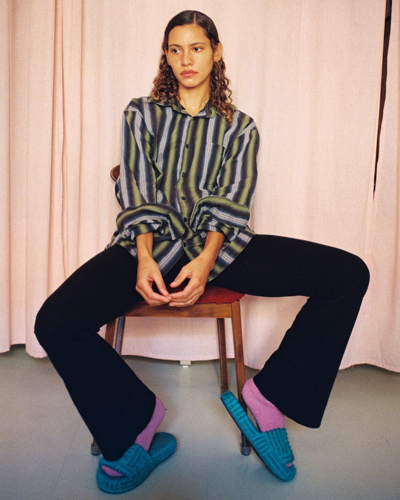 A woman sits in a chair wearing a striped shirt, black pants, pink socks, and teal sandals in an indoor setting with a pink backdrop.