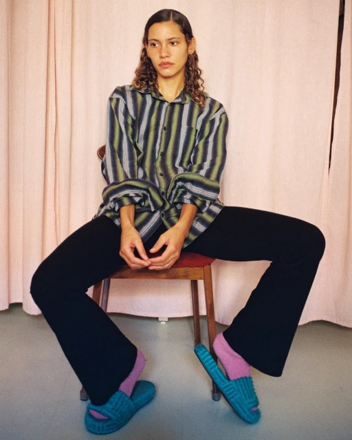 A woman sits in a chair wearing a striped shirt, black pants, pink socks, and teal sandals in an indoor setting with a pink backdrop.