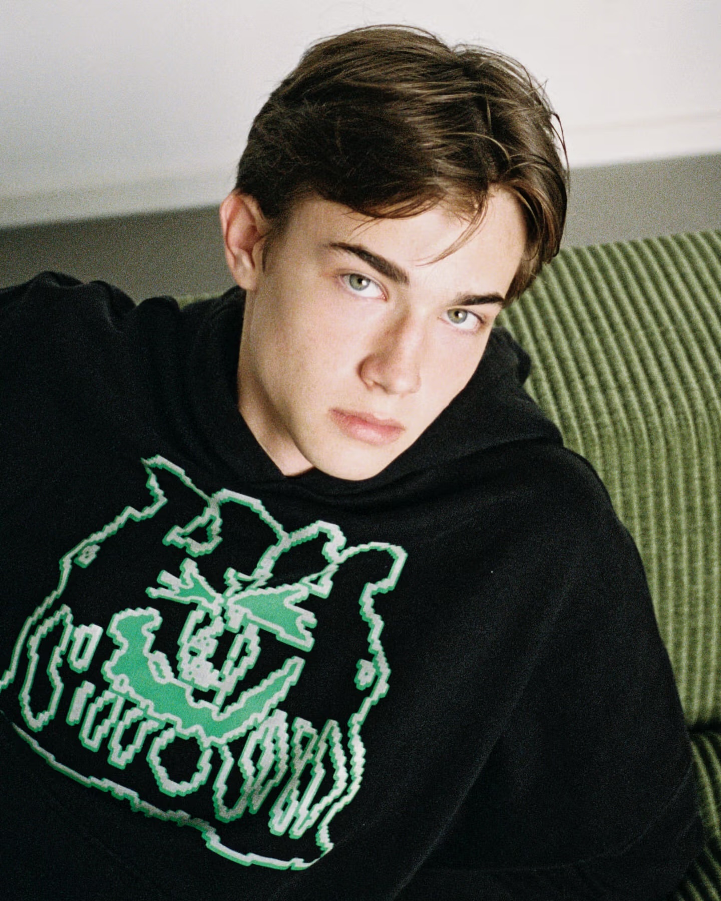 Portrait of a young adult male with brown hair, wearing a graphic black hoodie, looking directly at the camera in an indoor setting.
