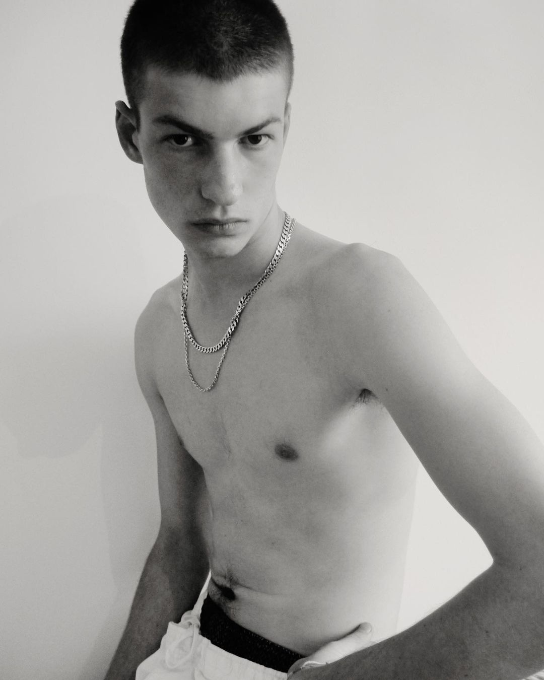 A shirtless young adult male with a closely cropped haircut poses indoors with a neutral expression, wearing layered silver necklaces against a plain background.
