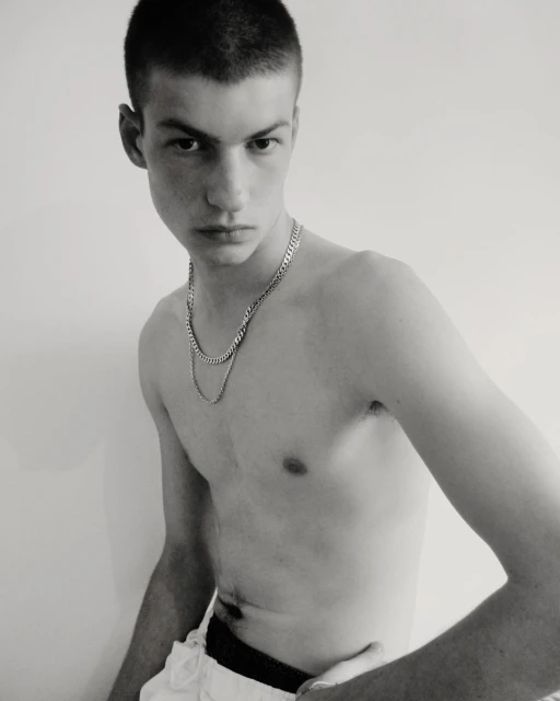 A shirtless young adult male with a closely cropped haircut poses indoors with a neutral expression, wearing layered silver necklaces against a plain background.