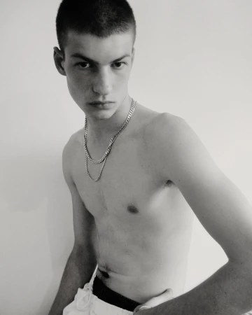A shirtless young adult male with a closely cropped haircut poses indoors with a neutral expression, wearing layered silver necklaces against a plain background.