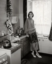 Monochrome portrait of a woman in a high-neck sweater leaning against a kitchen counter by Daniyel Lowden.