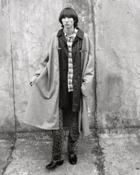 Man wearing a patterned trench coat and plaid shirt standing against a weathered concrete wall.