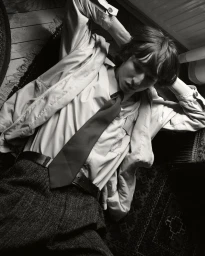 Monochrome portrait of a young man reclining with hands behind his head, wearing a collared shirt, tie, and textured trousers.