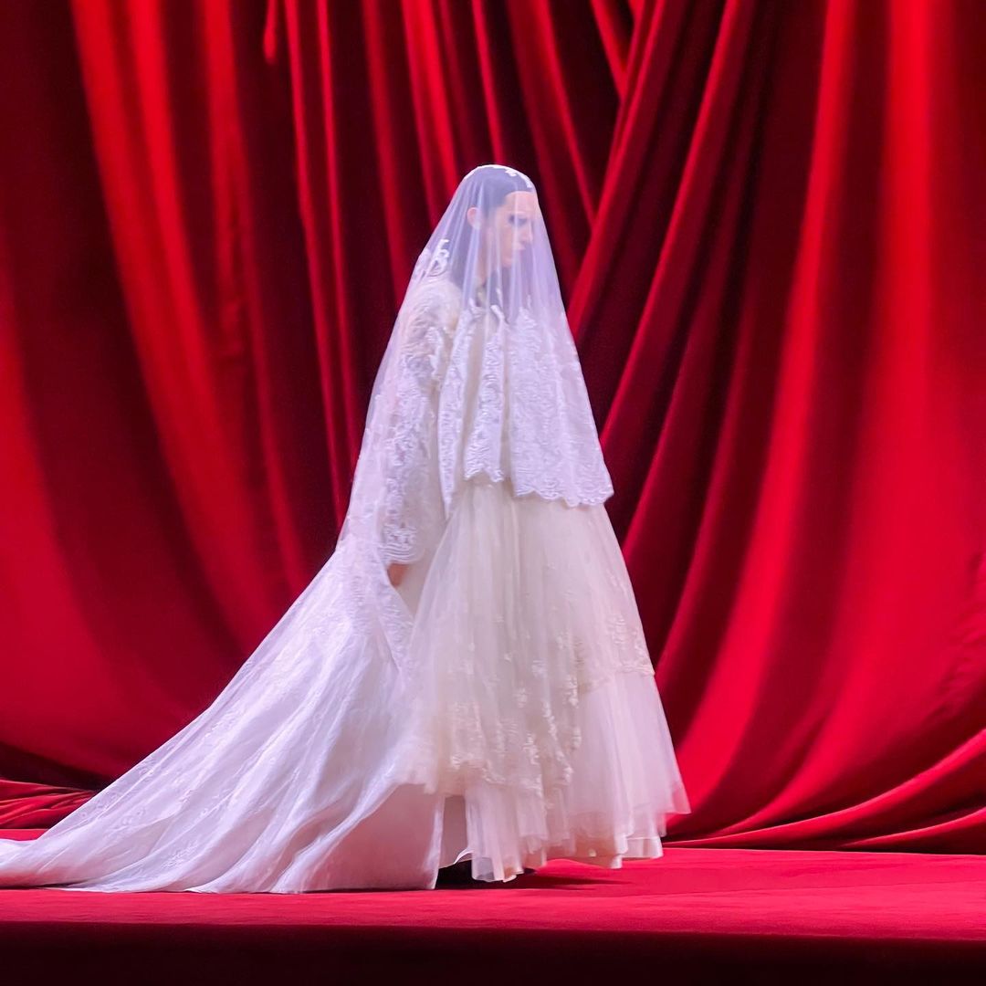 An adult female model showcases an off-white layered dress and veil on a runway in front of a red draped background in a fashion show.