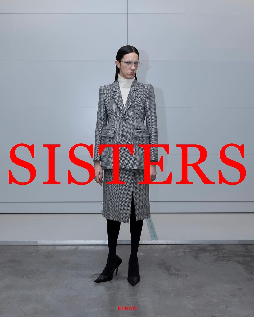 A woman in a grey patterned suit and heels stands against a pale grey wall, with the word 'SISTERS' in large red letters overlaying the image.