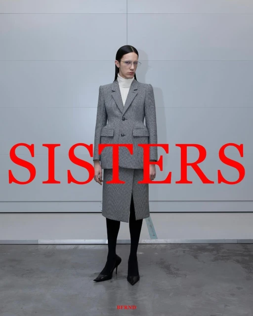 A woman in a grey patterned suit and heels stands against a pale grey wall, with the word 'SISTERS' in large red letters overlaying the image.