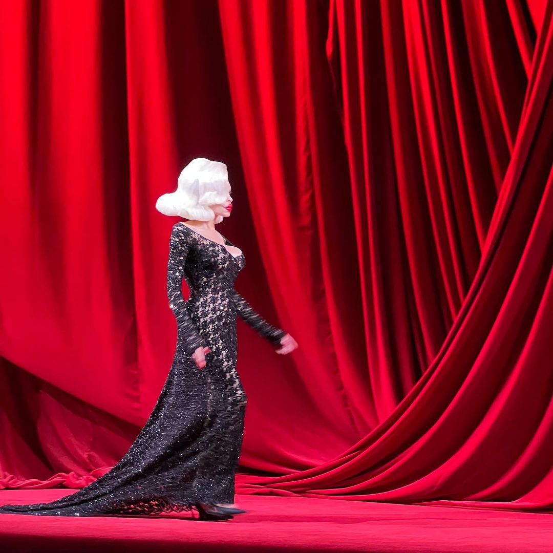 Fashion model walks the runway in a black lace gown against a red curtain backdrop, showcasing elegance and design in a stage setting.