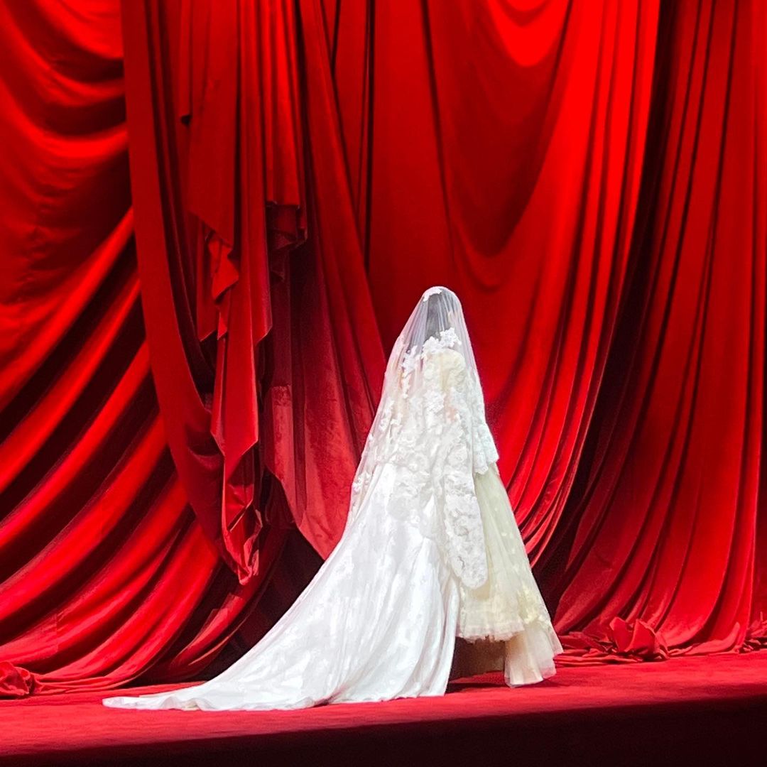 Back view of an unrecognizable person in a white wedding dress with lace details and a long veil, standing in front of red velvet curtains.