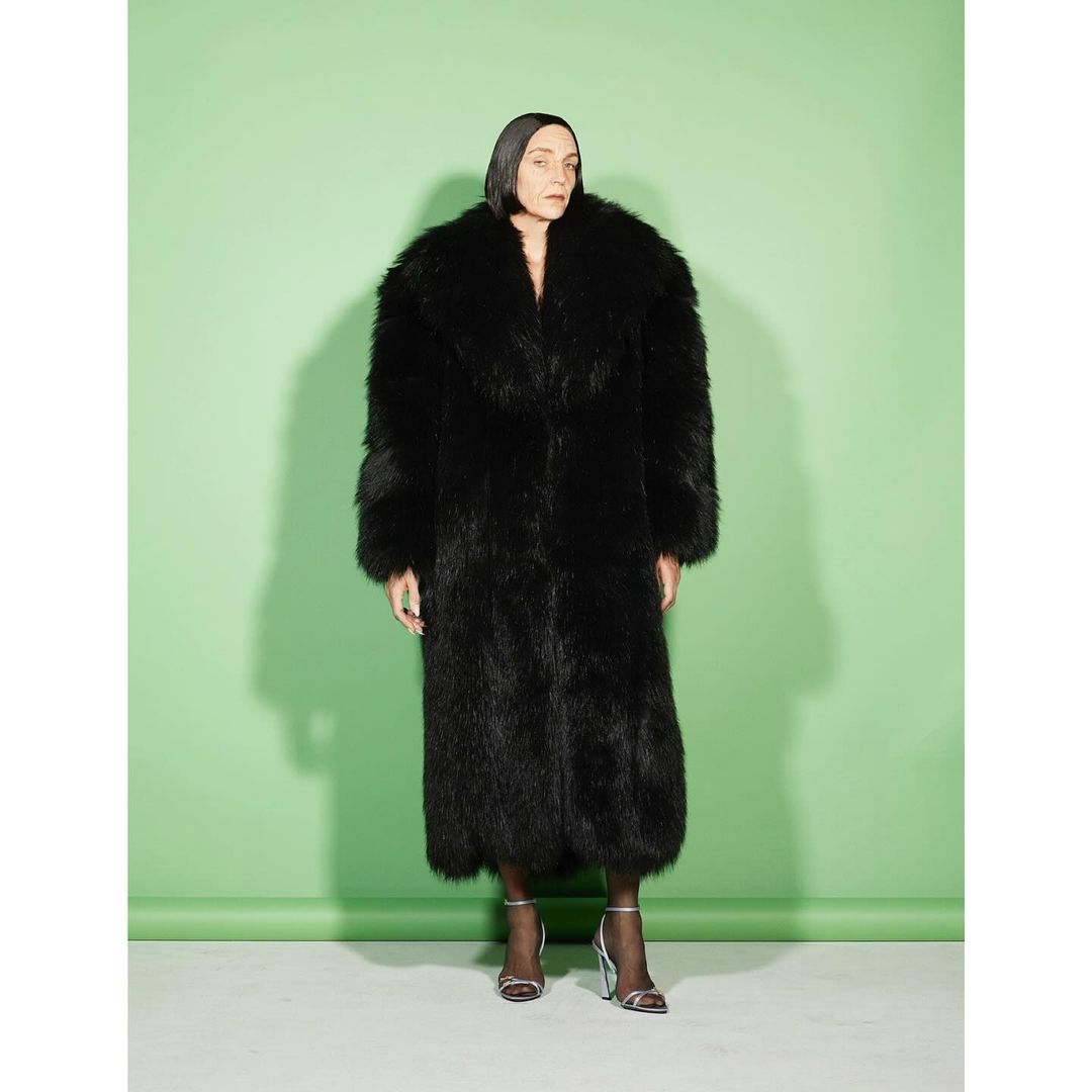 Adult woman wearing a long black fur coat and high heels poses against a light green background in a studio shot.