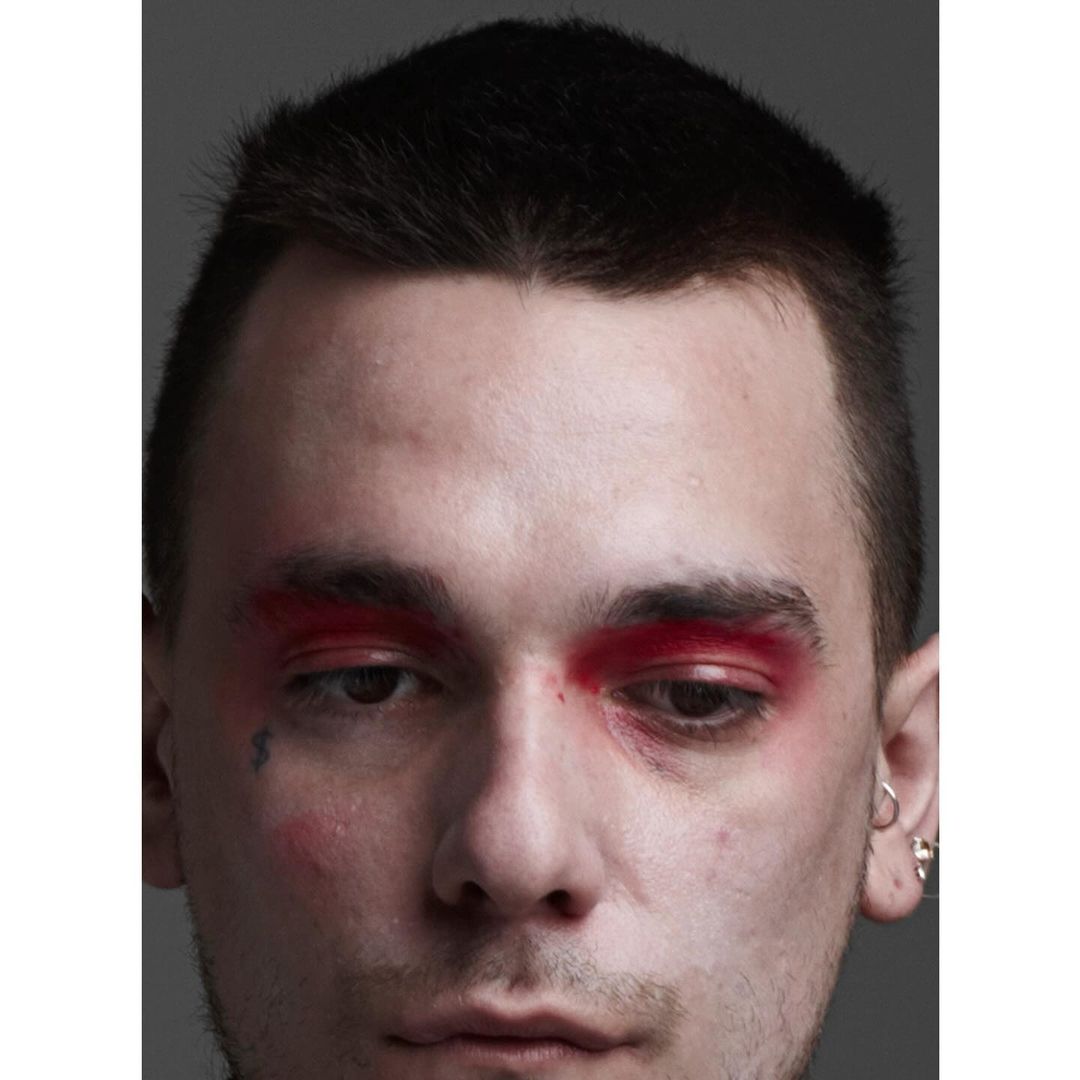 Portrait of a young adult male with dark hair and light skin, wearing vivid red eye makeup and multiple earrings, against a grey background.