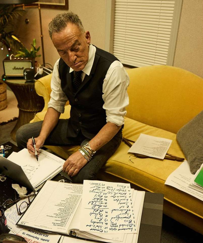 Bruce Springsteen on a yellow couch, writing on a paper with musical arrangements, wearing a black vest and grey jeans in an indoor environment.