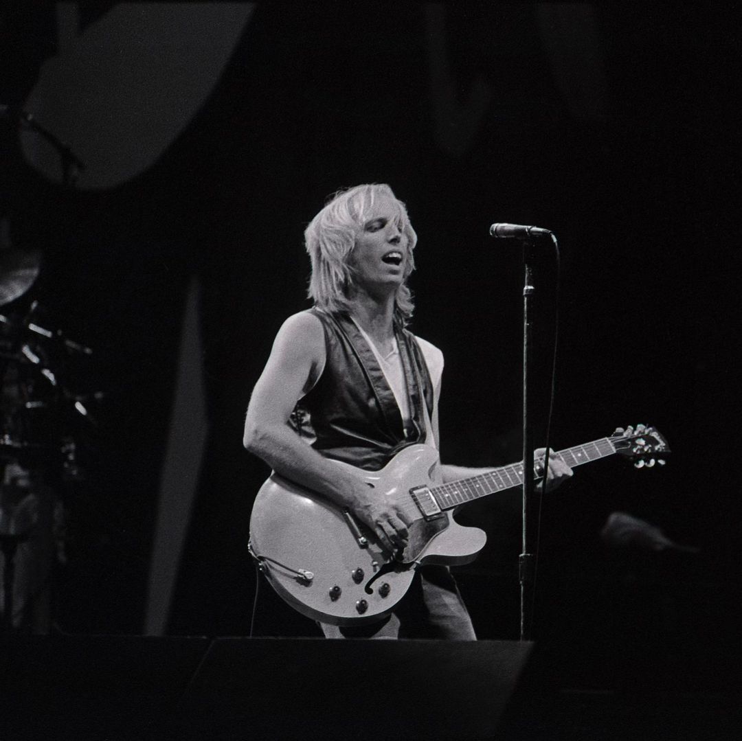 Monochrome photo of a fair-skinned adult male musician singing and playing an electric guitar on stage with a microphone.